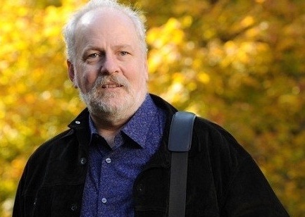Peter stands outside on a sunny day, with yellow autumn leaves in the background. He has a light skin tone and short, greying hair and beard. He is wearing a black jacket over a purple shirt.