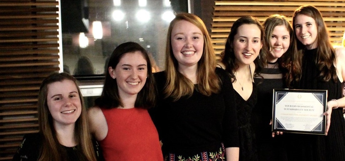 Six students stand together on a small stage under bright lights, smiling. They are wearing fancy outfits, and one is holding a framed certificate.