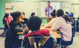 Group of students sitting around a table and looking at a man in a plaid shirt.. Group of students sitting around a table and looking at a man in a plaid shirt..