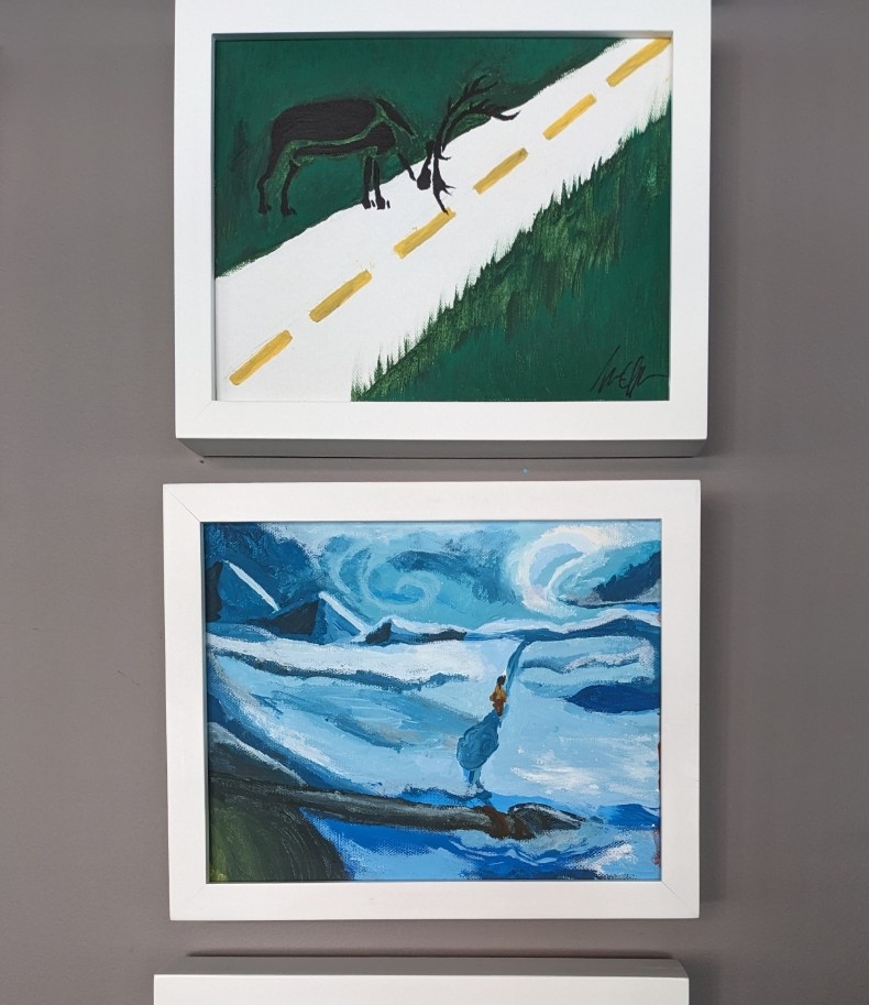 Two paintings hang on the wall: A stylized caribou examines a white paved road, and a small human figure walks through a swirling, snowy landscape.