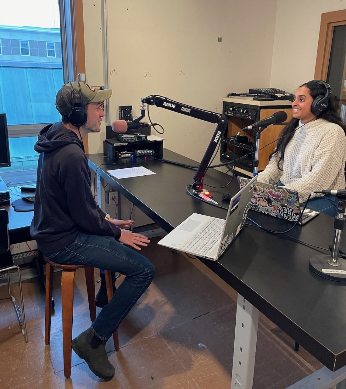 Two smiling students with headphones sit facing each other over a microphone in a recording studio.