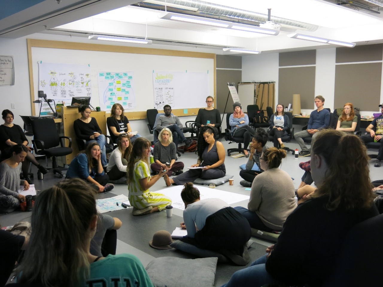 A group of people sit together in a large circle, with one person writing or drawing on a sheet of paper on the floor.