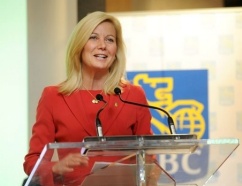 An woman with a light skin tone, blond hair and a red jacket makes an announcement in front of an RBC sign.
