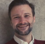 Andrew smiles. He has a light skin tone, short dark hair, and the beginnings of a beard and moustache.