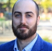 Gregory smiles for the camera. He has a light skin tone, dark brown hair with a receding hairline, and a neatly-trimmed beard and moustache.