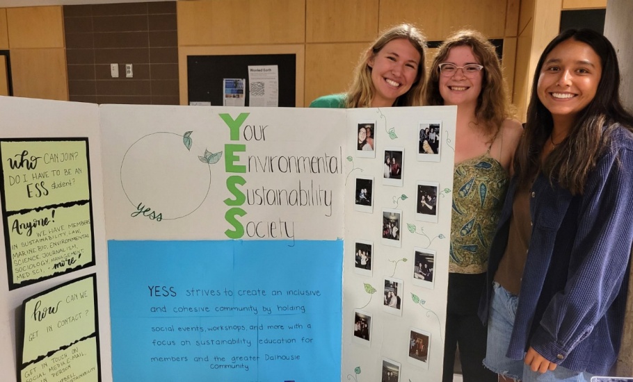 Three smiling students stand beside an information booth for Your Environmental Sustainability Society (YESS).