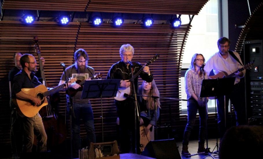 On a stage with purple spotlights shining from the back wall, students and faculty from the Sustainability String Band perform using guitars, banjos and percussion.