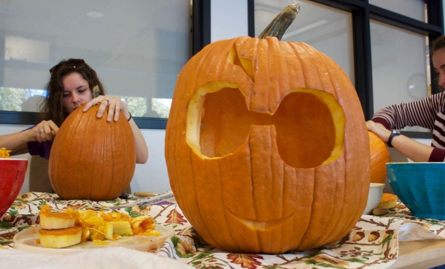 A carved pumpkin with big eyes and a tiny smile sits on a table, while students carve other pumpkins nearby.