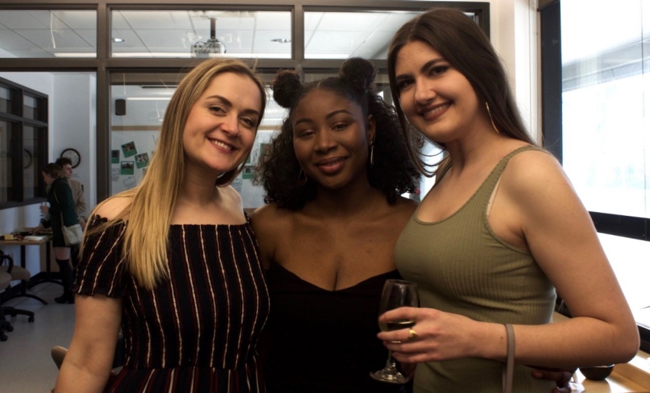 Three young women pose for a photo together, dressed in elegant outfits. One is carrying a wine glass.