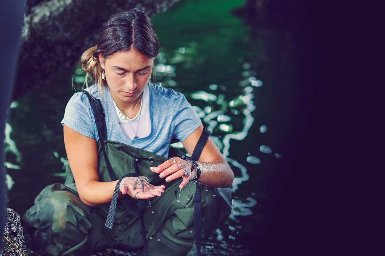 A student wearing waterproof overalls is crouching down by the dark shoreline, holding a small starfish in the palm of their hand.