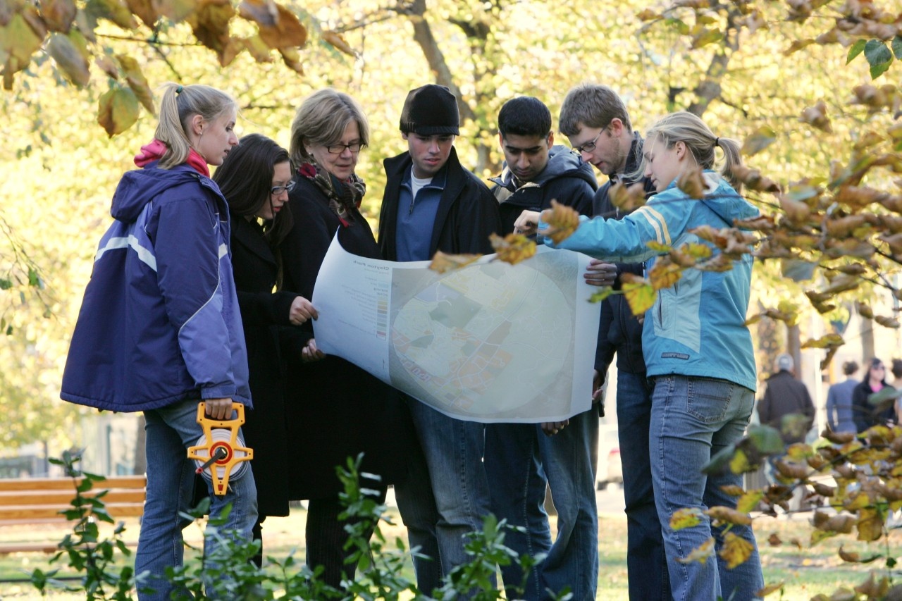 Outdoors, a small group of students in autumn gear gather around a map of campus with their instructor. One student is holding a reel of cable.