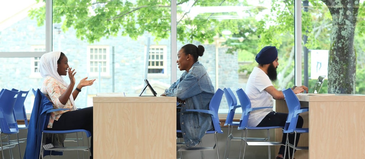 Two students converse while sitting on the concrete library sign outside the MacRae Library on a sunny day