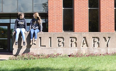 two students sit on sign outside MacRae library