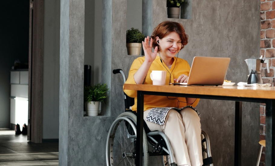 A woman in a wheelchair waves and smiles while looking at her laptop screen.