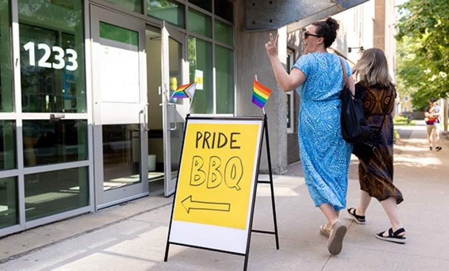 Two individuals walk into the pride BBQ beside a sign.