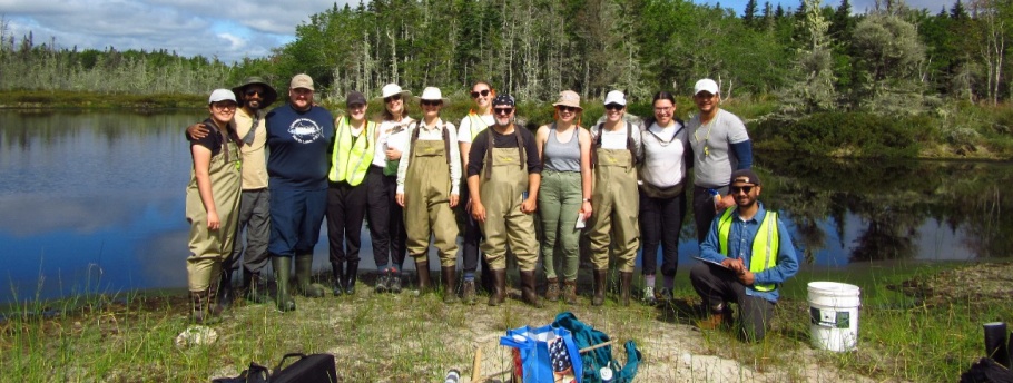 A group of MREM students dressed in winter gear stand in a forest looking at an instructor who is touching a tree